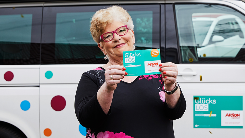 Gewinnerin Gaby steht lachend in einem Goldregen. Sie hält ein Glücks-Los in den Händen.