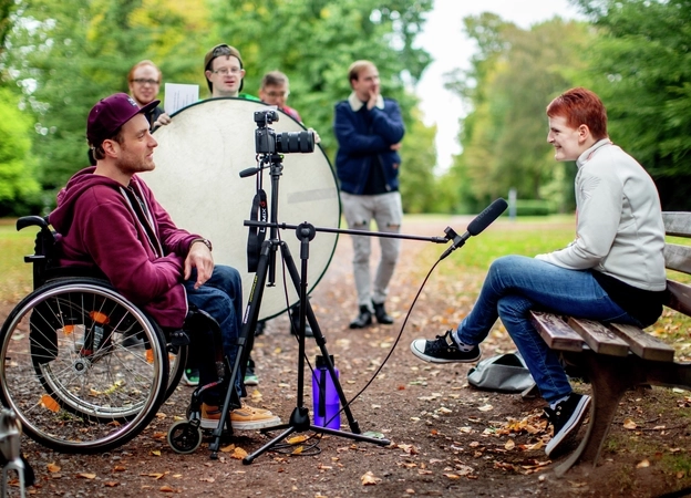 Ein junger Mann im Rollstuhl interviewt eine Frau in einem Park. Eine Kamera mit Stativ und Mikrofon sind auf die Frau gerichtet.