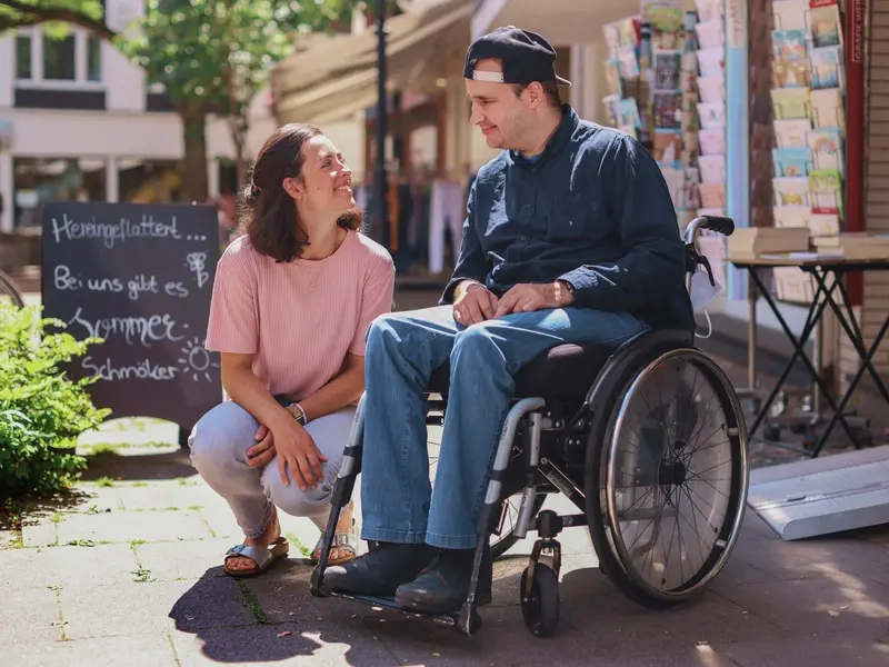 Ein Mann im Rollstuhl sitzt vor einer Buchhandlung. Neben ihm sitzt eine Frau in der Hocke. Die beiden lächeln sich an.