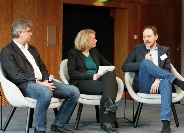 Auf einer Bühne sitzen drei Personen: Professor Dieter Katzenbach sitzt links, in der Mitte eine Moderatorin, Professor Hendrik Trescher sitzt rechts und spricht in ein Mikrofon. Rechts daneben steht eine Gebärdensprach-Dolmetscherin und übersetzt in Gebärdensprache.