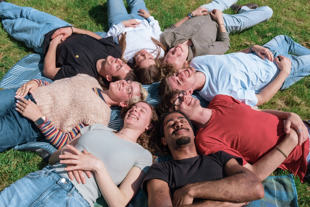 Eine diverse Gruppe von acht älteren Jugendlichen liegt sternförmig im Kreis angeordnet auf dem Rücken im Gras. Alle haben die Augen geschlossen und lachen.