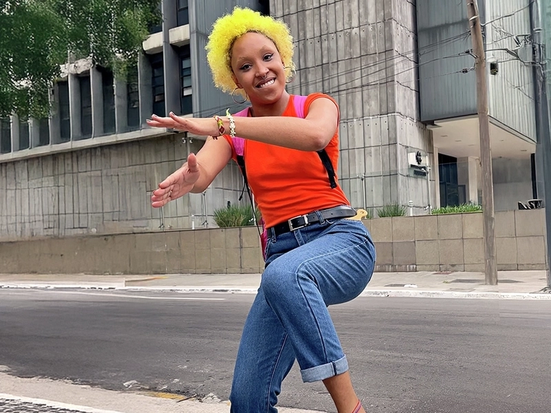 Eine junge Frau mit leuchtend gelben Locken tanzt fröhlich auf einer Straße in der Stadt. Sie trägt ein knallrotes T-Shirt, hochgekrempelte blaue Jeans und Sneaker. Ihr Gesicht strahlt Freude aus, während sie eine dynamische Tanzbewegung ausführt.