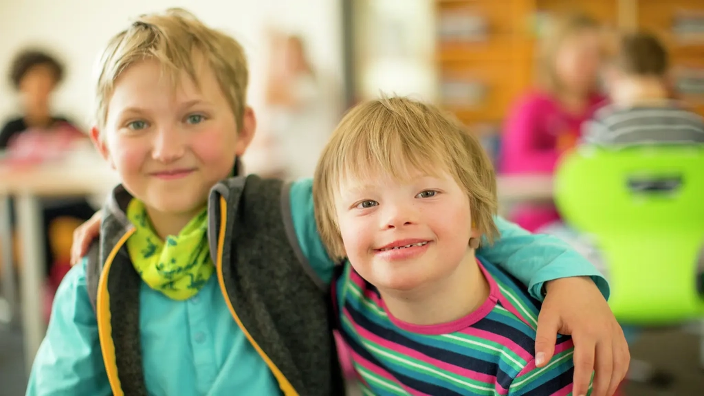 Zwei Jungen sitzen nebeneinander und lächeln in die Kamera. Einer der Jungen hat eine Behinderung, der andere legt seinen Arm um ihn.