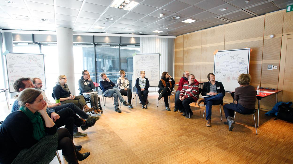 Teilnehmer sitzen in einem Stuhlkreis und betrachten ein selbstgestaltetes Plakat