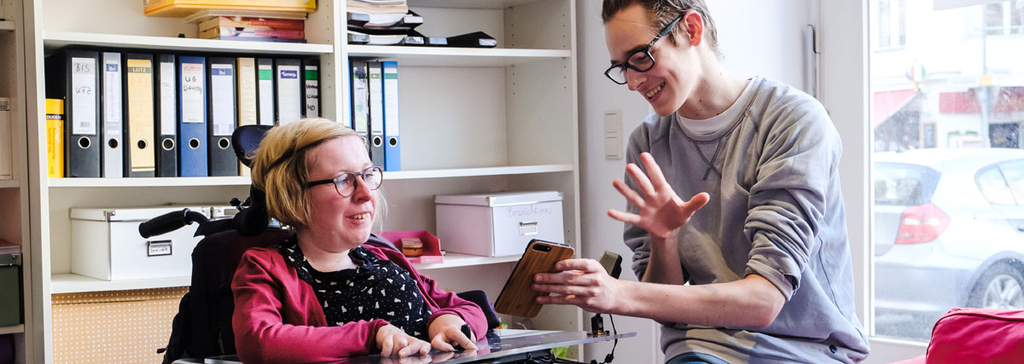 Eine Frau im Rollstuhl und ein Mann schauen auf ein Smartphone