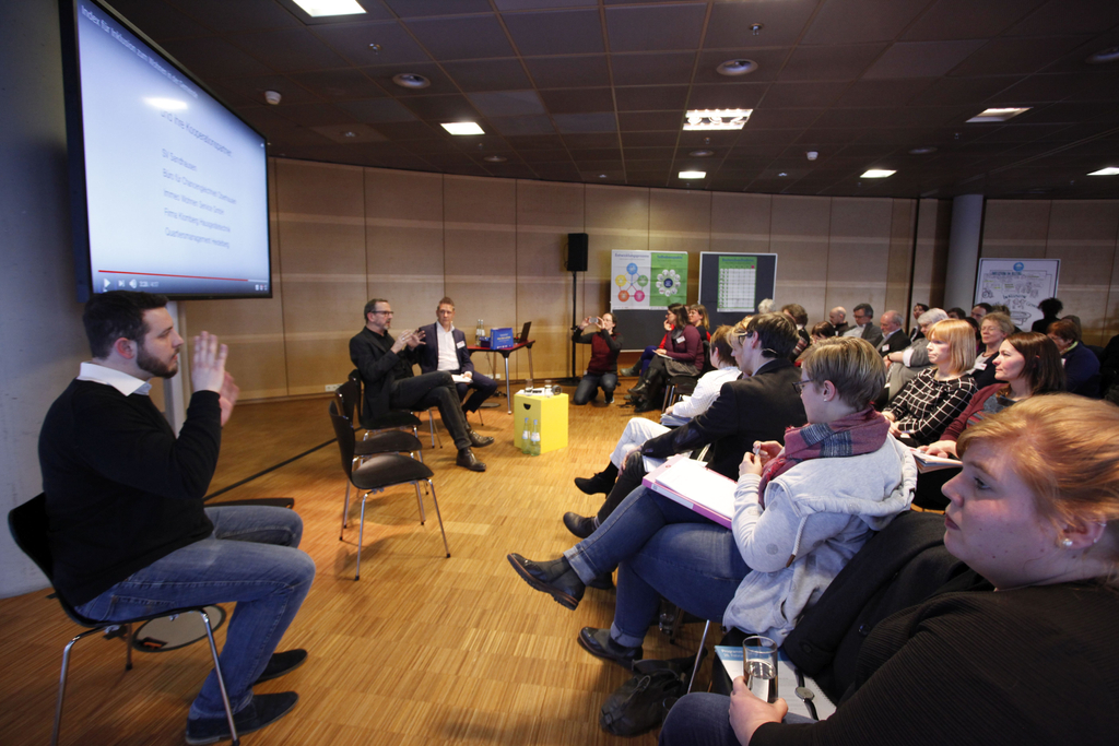 Der Referent Ulrich Niehoff erklärt den Teilnehmern etwas. Neben ihm sitzt der Moderator Stefan Burkhardt. Ein Gebärdensprachdolmetscher übersetzt für das Publikum.
