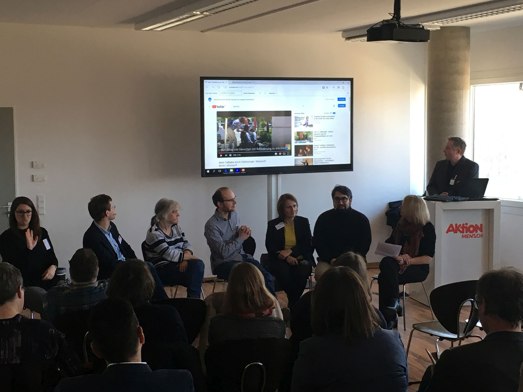 Die Teilnehmer und die Moderatorin Katja Nellissen schauen auf einen großen Bildschirm an der Wand. Dort wird ein youtube Video gezeigt. 