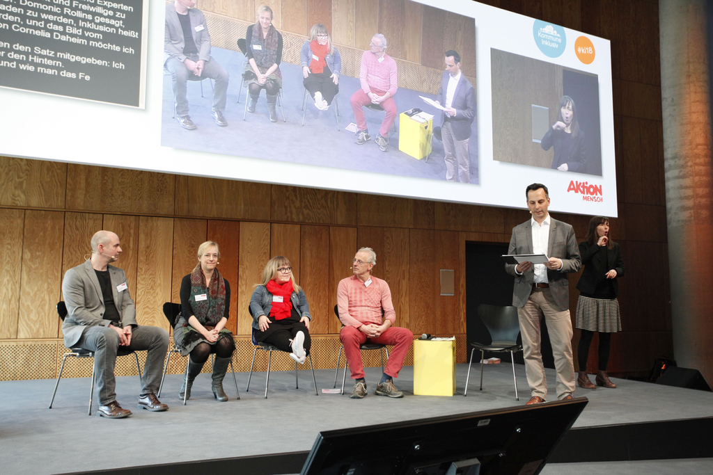 Der Moderator Frank Liffers steht auf der Bühne. Neben ihm sitzen Gäste.