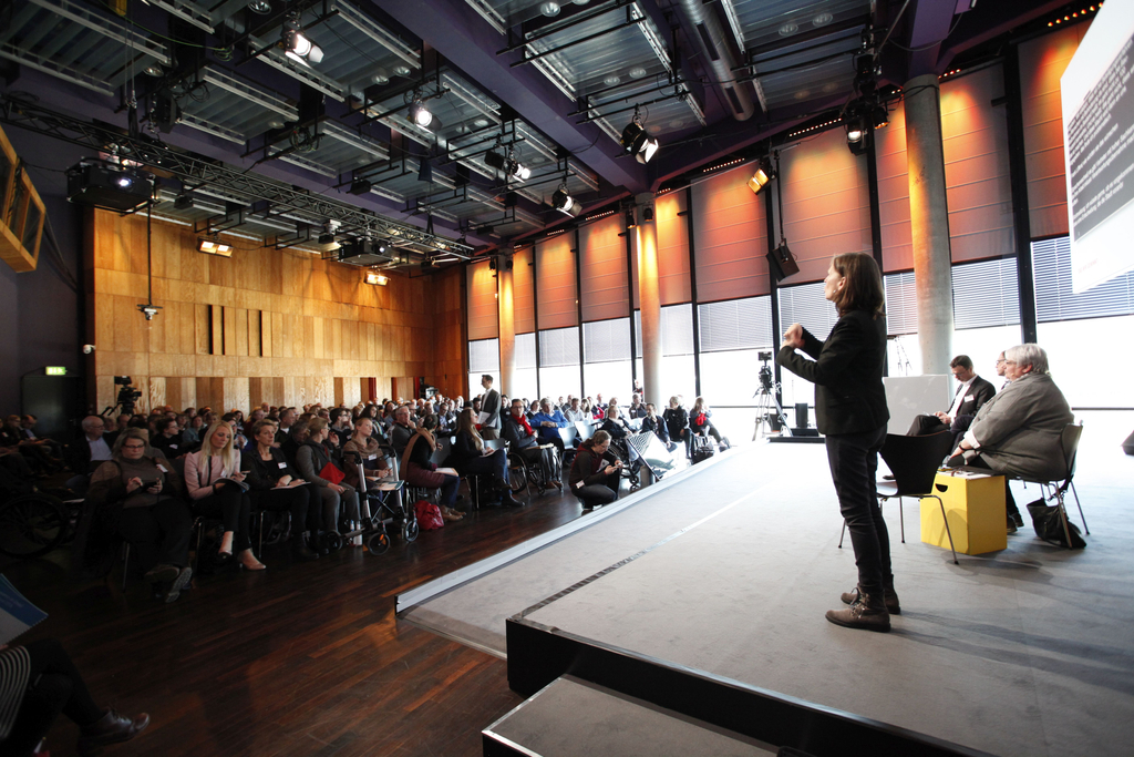 Der Moderator Frank Liffers steht mitten im Publikum. Eine Gebärdensprachdolmetscherin übersetzt