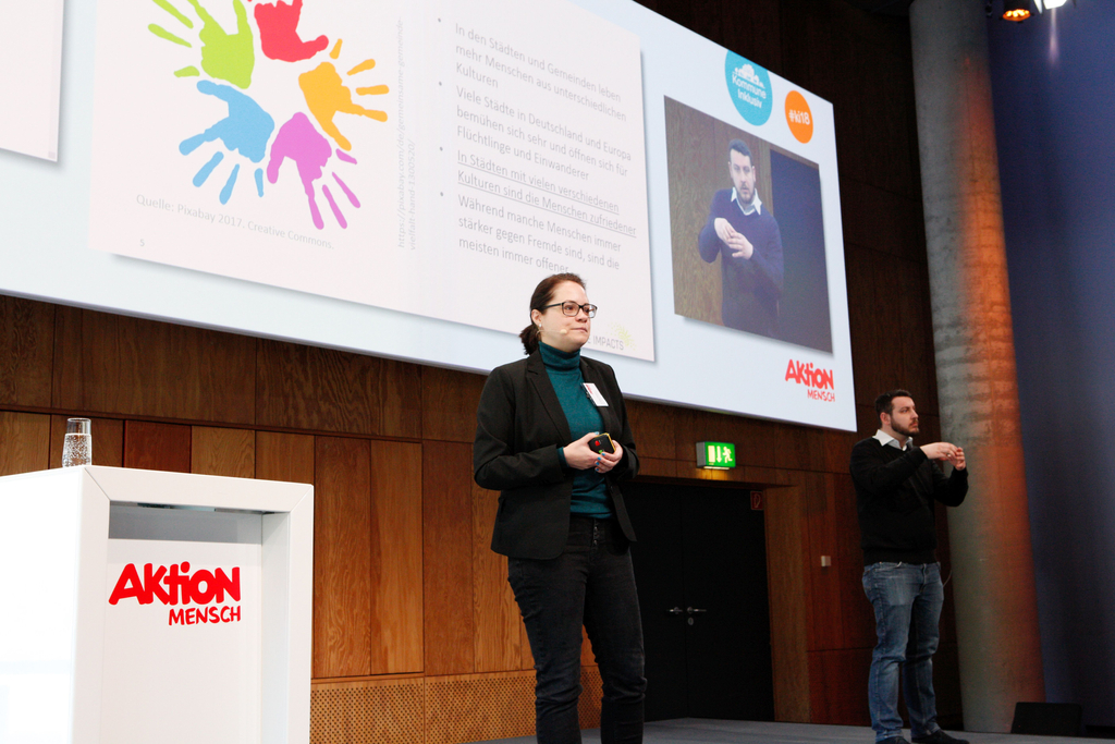 Cornelia Daheim auf der Bühne. Auf einer großen Leinwand hinter ihr wird der Vortrag übertragen. Ein Gebärdensprachdolmetscher übersetzt