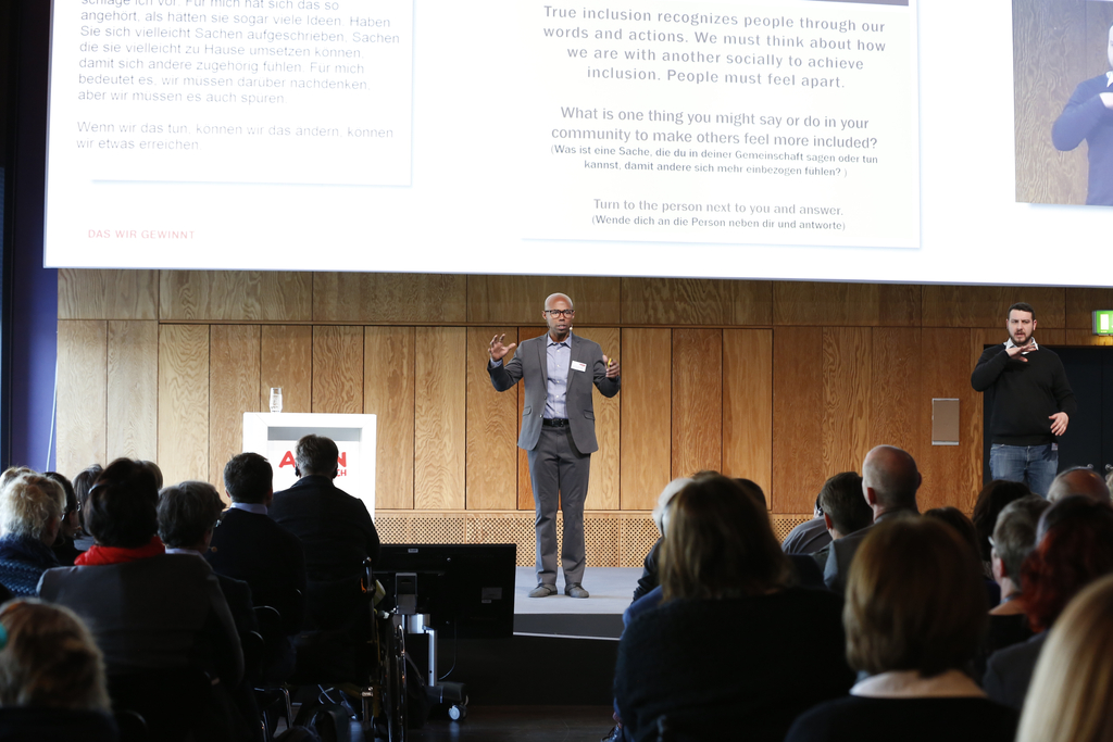 Domonic Rollins, PhD, bei seinem Vortrag auf der Bühne. Ein Gebärdensprachdolmetscher übersetzt