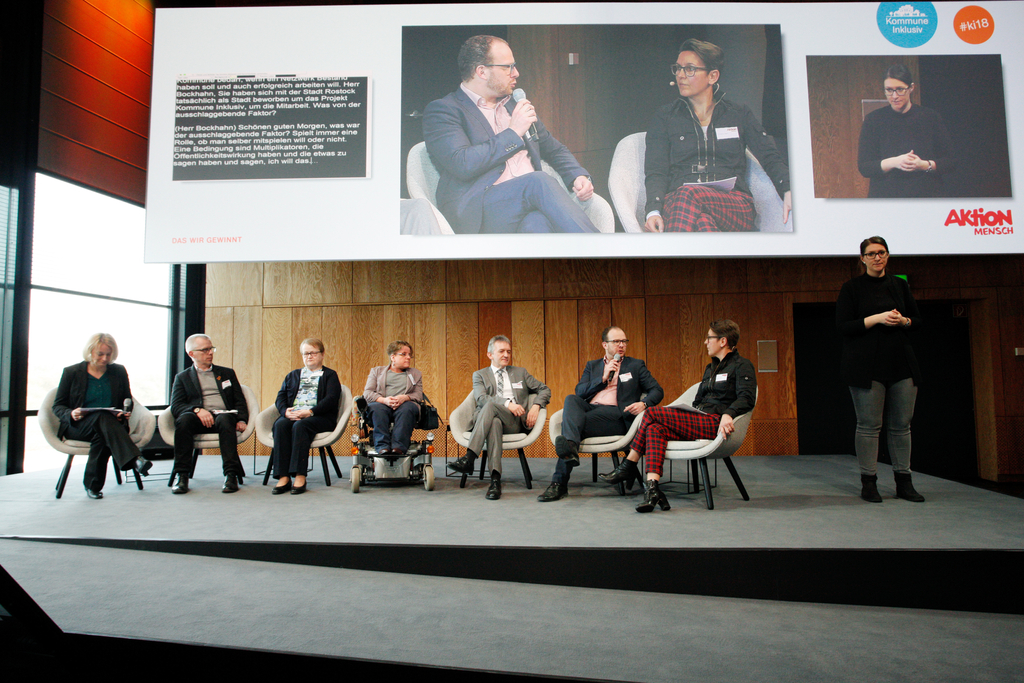 Auf der Bühne: Die Moderatorin Katja Nellissen mit Johannes Blaurock, Elisabeth Paulus, Gracia Schade, Gerhard Suder, Steffen Bockhahn und Christina Marx. Eine große Leinwand überträgt alles im Hintergrund. Eine Gebärdensprachdolmetscherin übersetzt