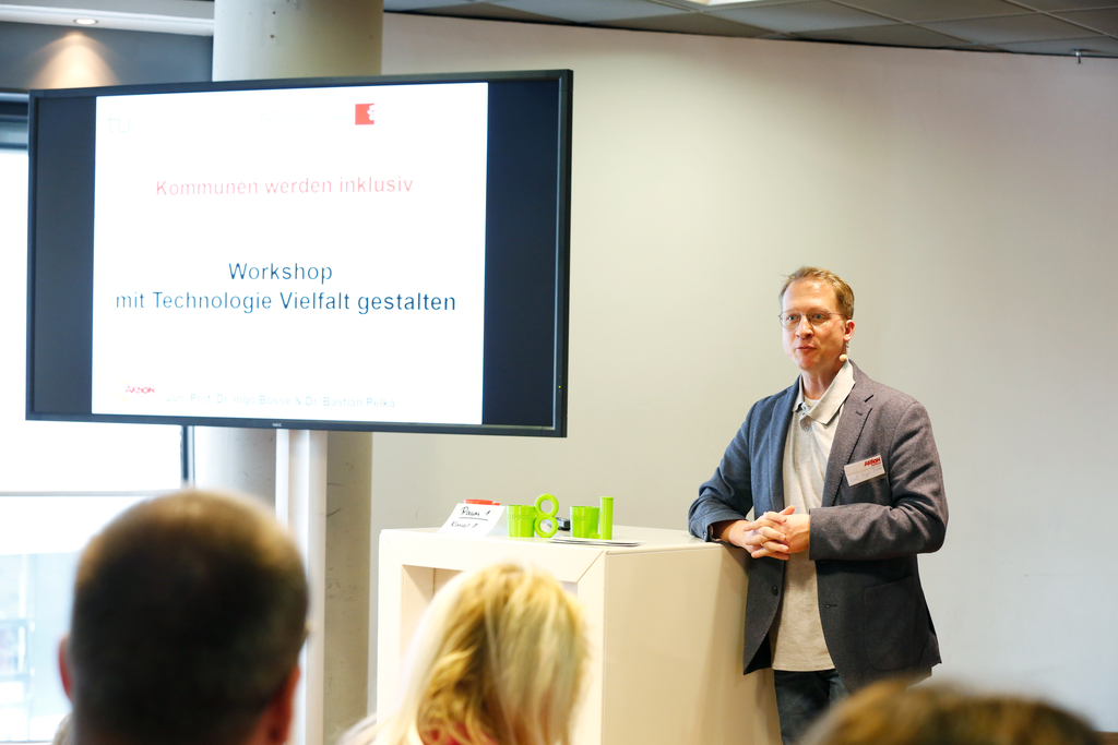 Der Referent Ingo Bosse steht neben einem Pult auf dem 2 Tassen stehen, die mit dem 3D Drucker gemacht wurden