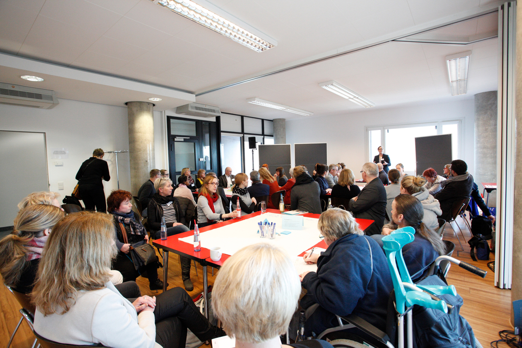 Teilnehmer sitzen um große Tische verteilt. Die Referentin Marion Theisen erklärt etwas