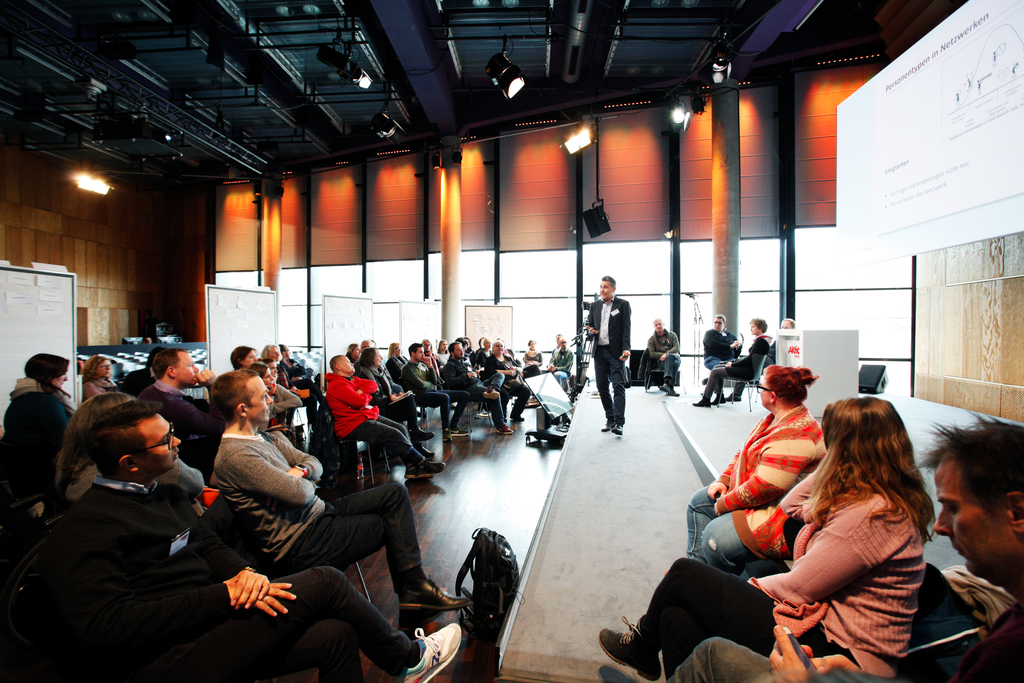 Der Referent Stefan Burkhardt steht auf der Bühne. Viele Teilnehmer hören ihm zu