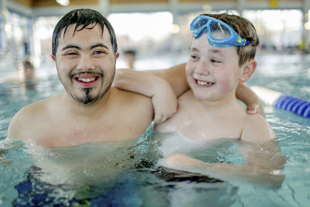 Zwei Jugendliche sind zusammen in einem Schwimmbecken. Einer der beiden Jungen hat eine Behinderung, der andere nicht. Er trägt eine Schwimmbrille auf dem Kopf.