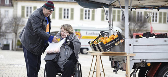 Mann reicht einer Frau im Rollstuhl eine Apfelsine