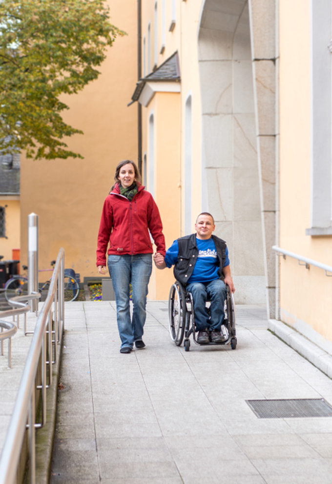 Ein Mann im Rollstuhl und eine Frau Hand in Hand auf einer Rampe vor einem Gebäude