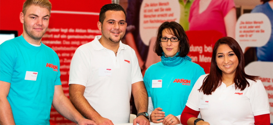 Lächelnde Mitarbeiter und Mitarbeiterinnen der Aktion Mensch vor dem Infostand der Aktion Mensch