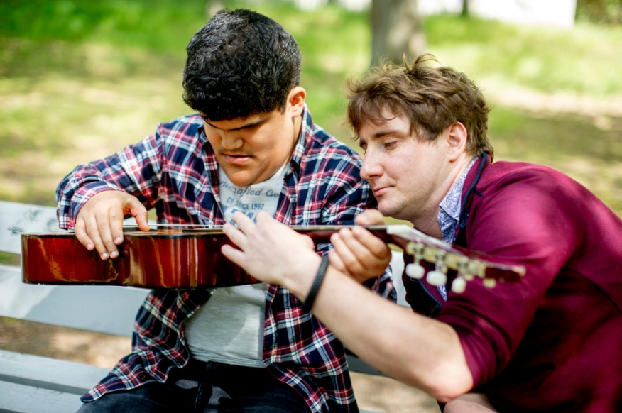 Zwei junge Männer mit und ohne Behinderung sitzen auf einer Parkbank und üben gemeinsam Gitarrespielen.