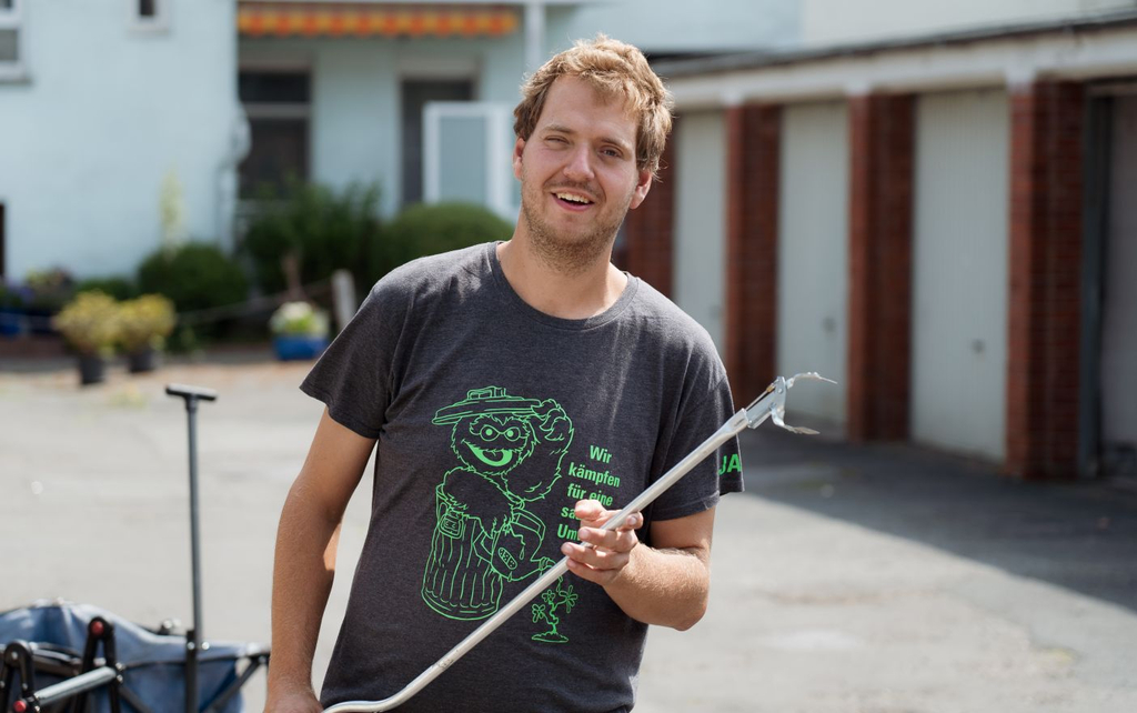 Ein junger Mann mit blonden Haaren steht mit einer Greifzange in der Hand auf einem Hof vor einer Reihe Garagen. Er trägt ein graues T-Shirt auf dem eine Comicfigur abgebildet ist.