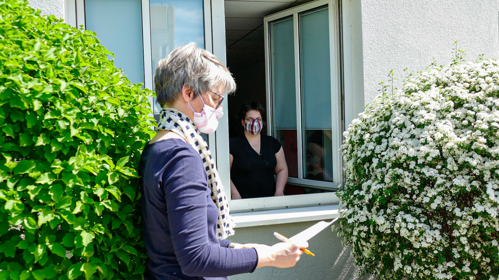Eine Frau mit Mundschutz steht mit einem Klemmbrett an einem Fenster. Im Fenster steht eine weitere Frau, auch mit Mundschutz.