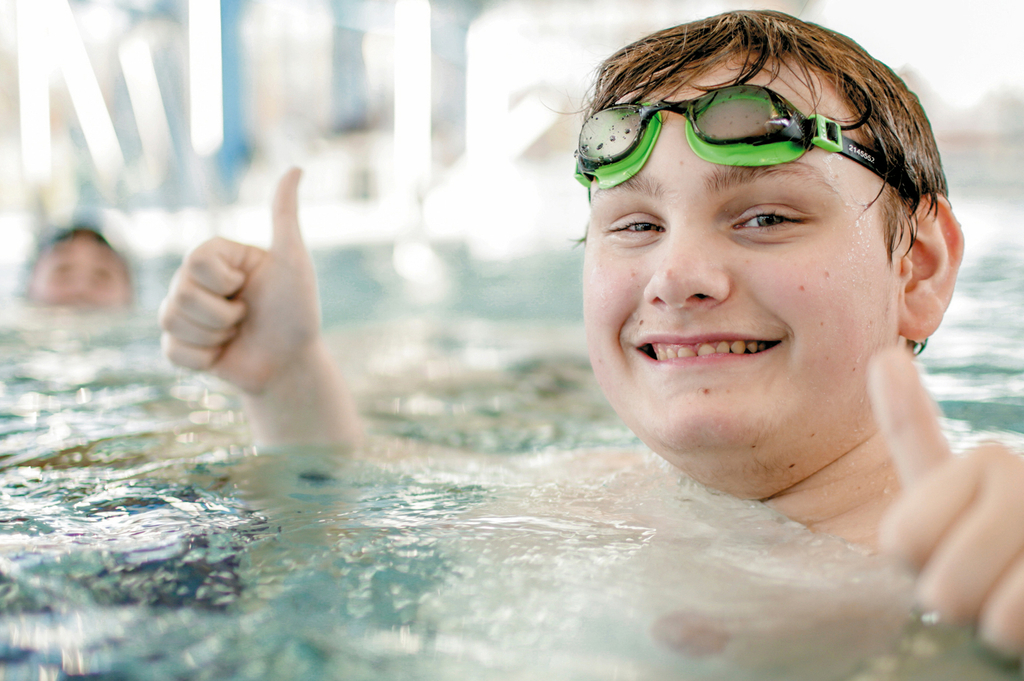 Ein Junge im Schwimmbecken lächelt in die Kamera und hebt die Daumen.