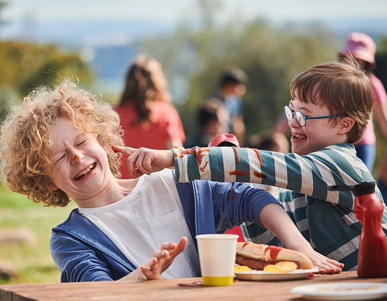 Zwei Jungen mit und ohne Behinderung sitzen an einem Tisch im Freien und spielen mit Ketchup.