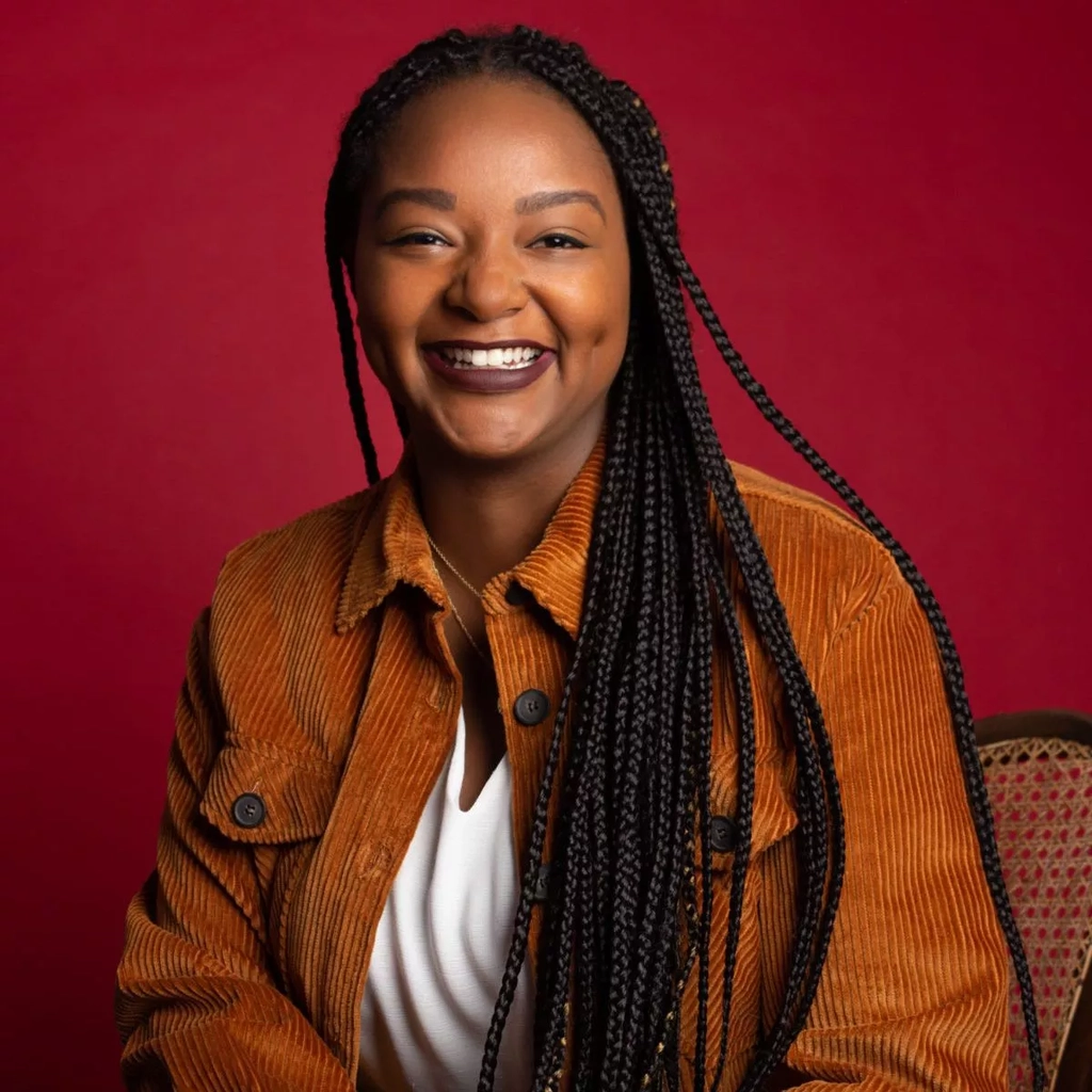 Eine junge Frau mit vielen schwarzen geflochtenen Strähnen sitzt lächelnd vor einer dunkelroten Wand.