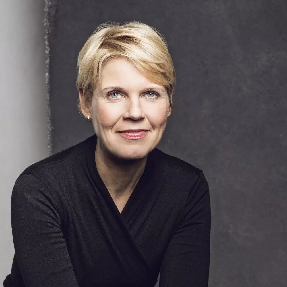 Eine Frau mit kurzen blonden Haaren sitzt vor einer grauen Wand und lächelt freundlich in die Kamera.