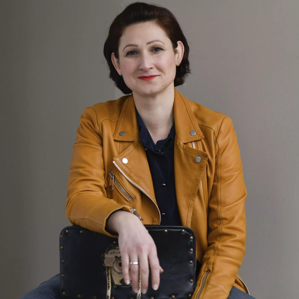 Eine junge Frau mit schwarzen, mittellangen Haaren sitzt rittlings auf einem schwarzen Lederstuhl. Sie trägt eine helle Lederjacke.