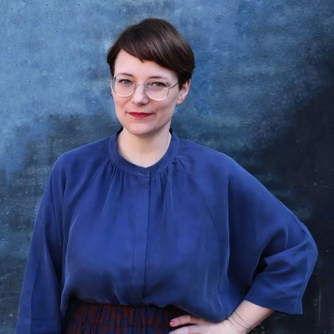 Eine junge Frau mit kurzen dunklen Haaren und Brille steht vor einer blauen Wand. Sie trägt eine lockere blaue Bluse und stützt einen Arm in die Hüfte.