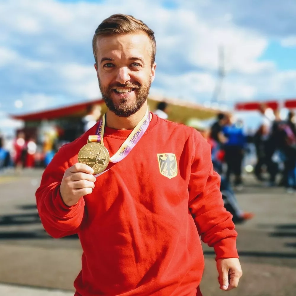 Ein kleinwüchsiger Mann trägt einen roten Pullover der deutschen Nationalmannschaft und hält seine Goldmedaille in die Kamera.