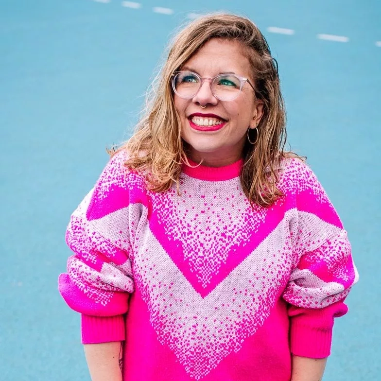 Eine junge Frau mit Brille und pinkem Pullover steht vor einem hellblauen Hintergrund.