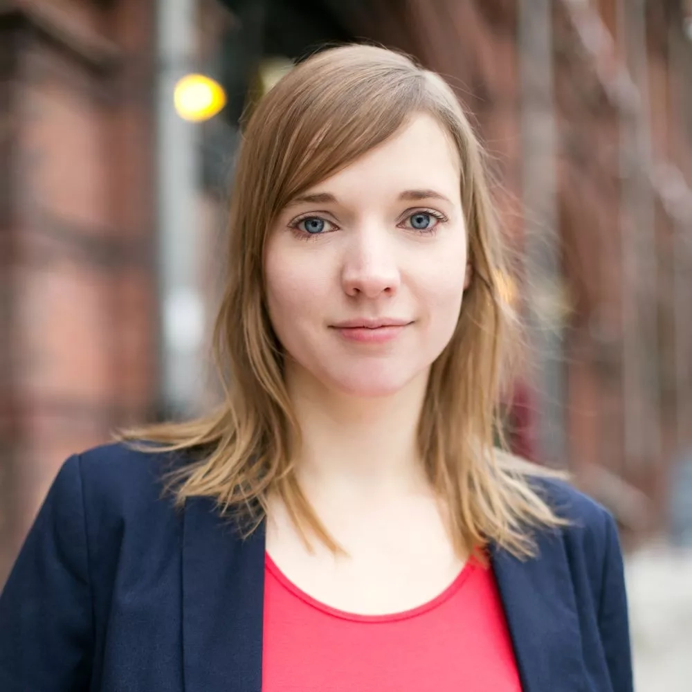 Eine junge Frau mit dunkelblondem Haar steht vor einem Backsteingebäude. Sie trägt ein rotes T-Shirt und einen dunklen Blazer.