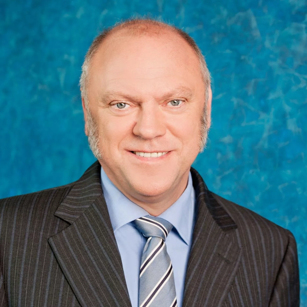 Ein Mann mit Halbglatze steht vor einer blau gemusterten Wand. Erträgt einen gestreiften dunklen Anzug und eine Krawatte.