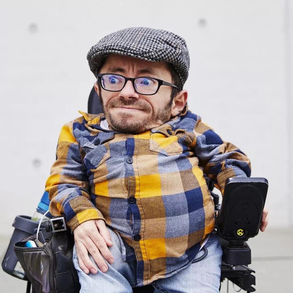 Ein junger Mann sitzt im Rollstuhl. Er trägt ein gelb-grau kariertes Hemd, eine Brille und eine Schirmmütze.