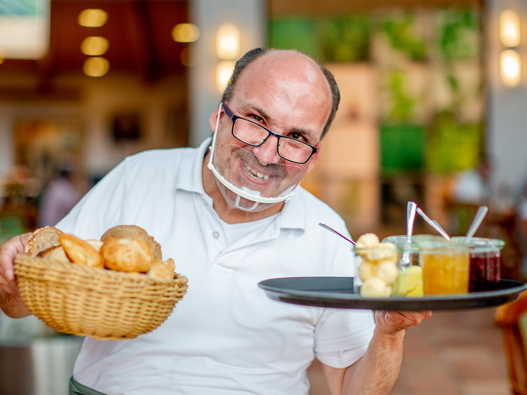 Ein Mann mit durchsichtigem Mundschutz halt ein Tablet mit Marmelade und ein Korb mit Brötchen in den Händen.