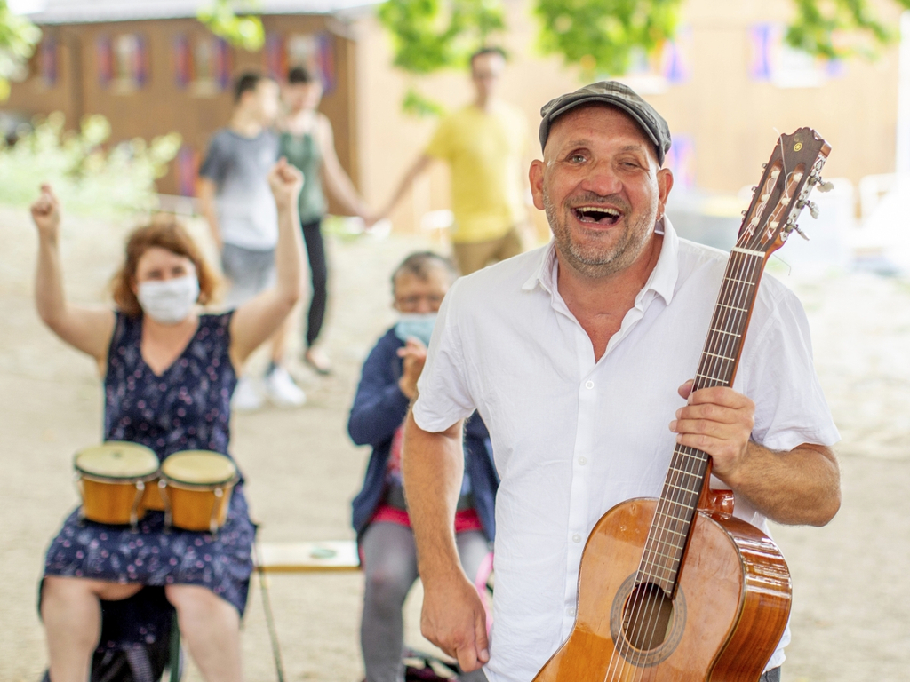 Ein Mann mit Gitarre lacht in die Kamera. Hinter ihm spielt eine Frau auf einer Trommel.