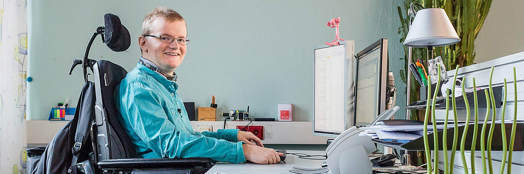 Ein junger Mann sitzt lächelnd in seinem Rollstuhl vor einem Computerbildschirm.