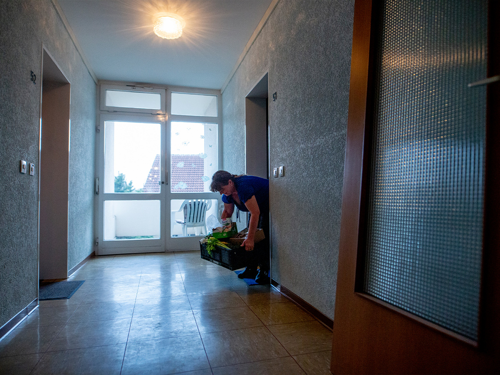 Eine Frau hebt die Lebensmittelkiste vor ihrer Wohnungstür hoch.
