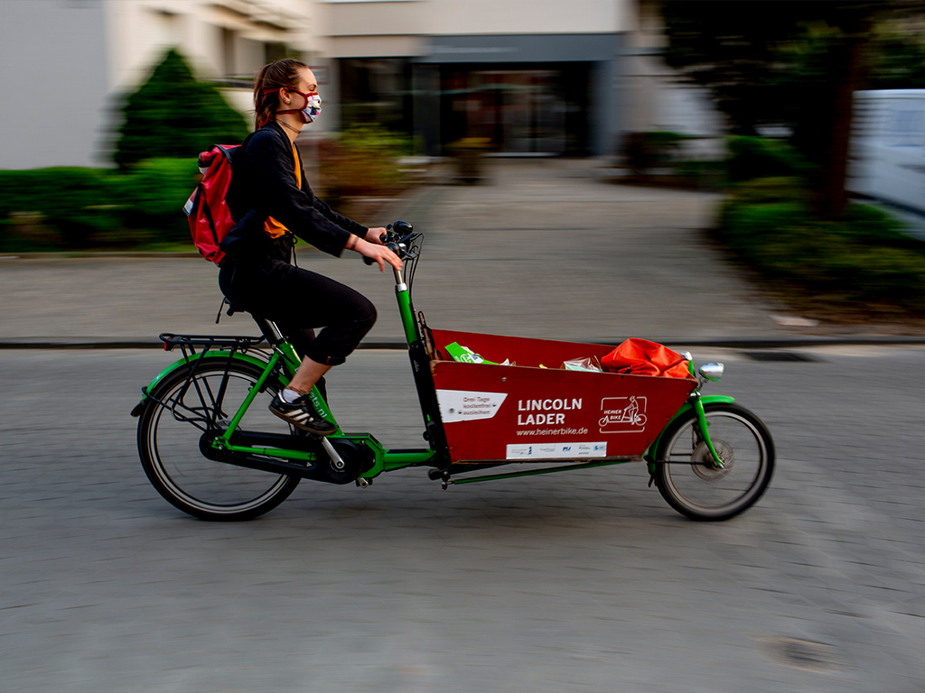 Ein junge Frau mit Atemschutzmaske fährt auf einem Lastenrad.