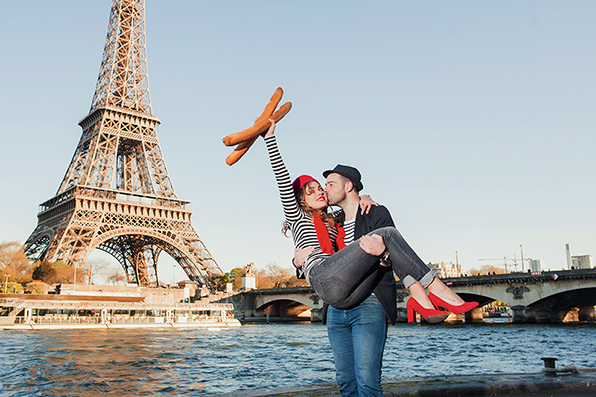 Ein junger Mann trägt eine junge Frau auf seinen Armen. Sie hält zwei Baguette-Brote in der Hand. Im Hintergrund sieht man den Eiffelturm.