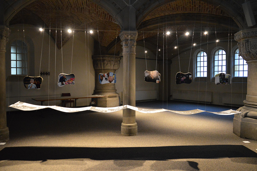 In einem Raum mit gotischen Säulen hängen von der Decke verschiedene Bildausschnitte herab. Darunter hängt ein langer Papierbogen.