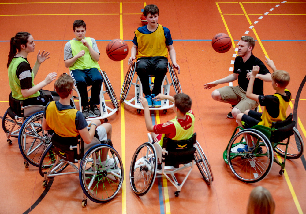 Jugendliche Rollstuhlbasketballer bilden in einer Turnhalle eine Kreis und werfen sich die Bälle zu