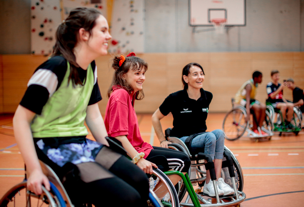 Drei Mädchen sitzen in Rollstühlen und spielen Rollstuhlbasketball
