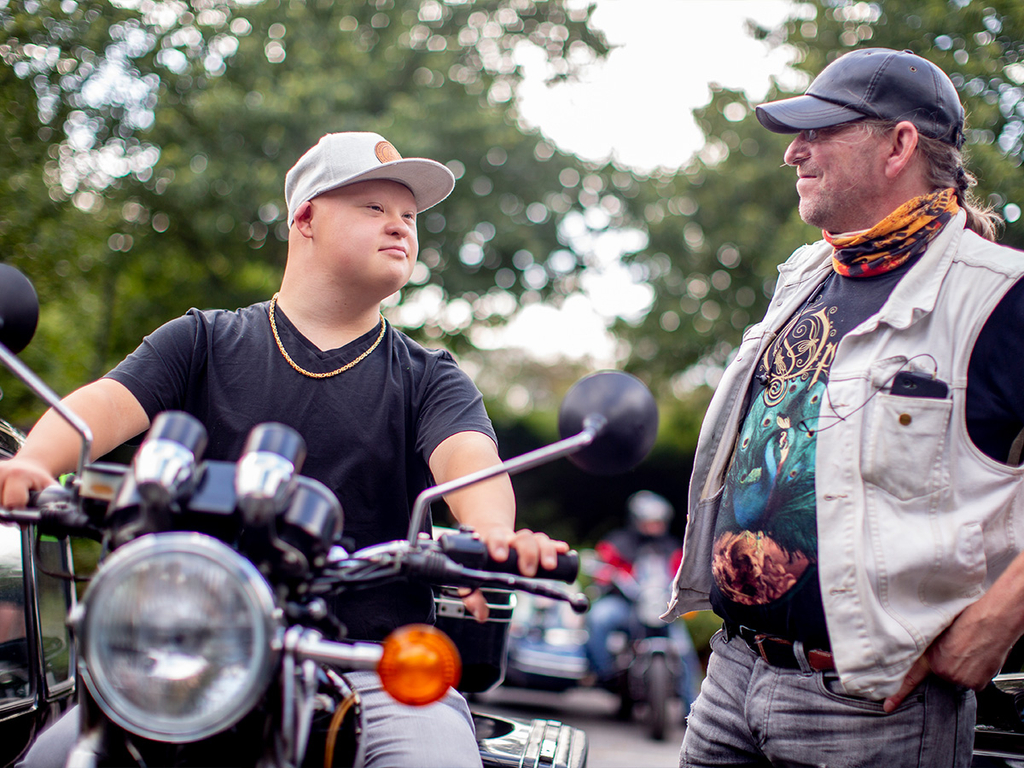 Zwei Männer unterhalten sich. Der linke Mann sitzt auf einem Motorrad.