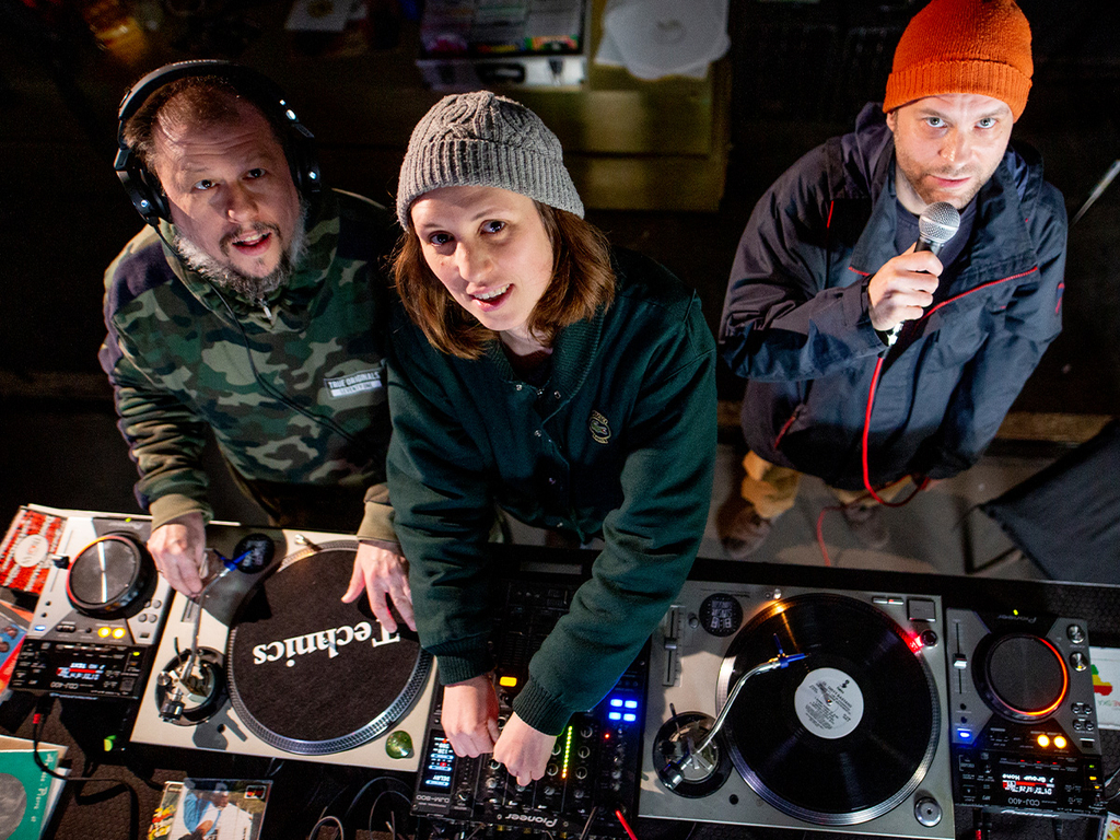 Draufsicht auf das DJ-Pult. Drei Dj*s schauen nach oben in die Kamera.