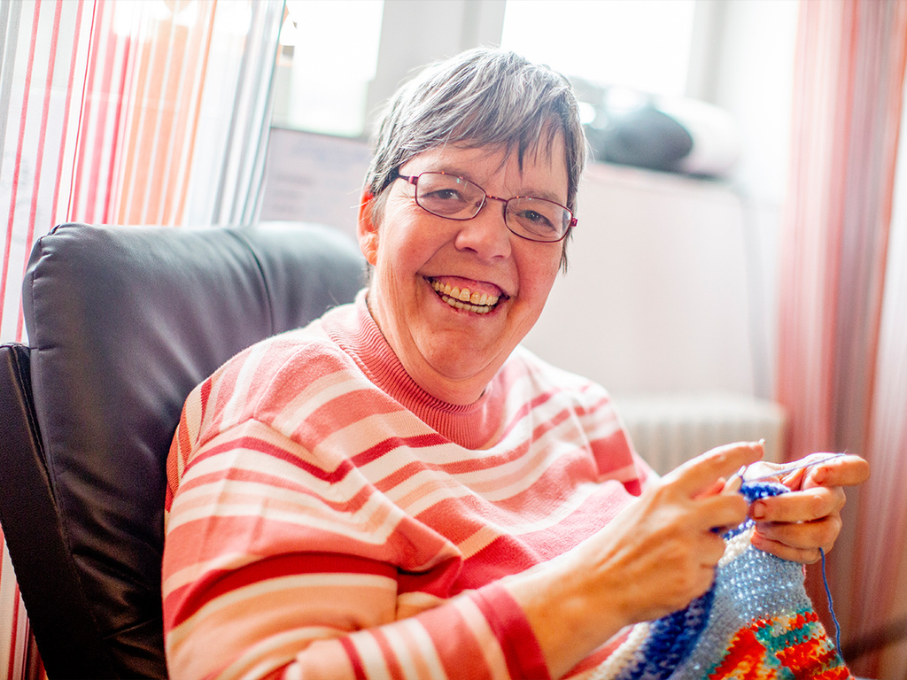 Eine Frau strickt und lacht in die Kamera.