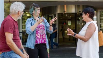 Zwei Frauen unterhalten sich in Gebärdensprache und ein Mann ischaut zu. 